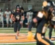 A football player in a black uniform runs with the ball during a night game, showcasing dynamic action on the field.