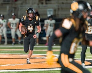 A football player in a black uniform runs with the ball during a night game, showcasing dynamic action on the field.