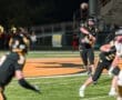 Quarterback throws a pass during a night football game, with teammates in black uniforms on a lit field.