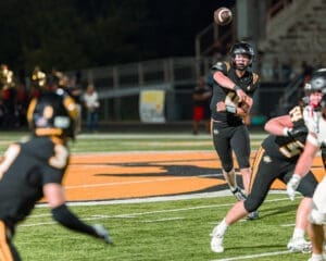 Quarterback throws a pass during a night football game, with teammates in black uniforms on a lit field.