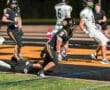 High school football player in black uniform runs towards the end zone with the ball during an intense game night.