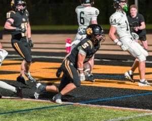 High school football player in black uniform runs towards the end zone with the ball during an intense game night.