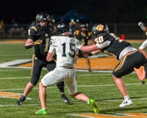 Two football players tackle an opposing player in a nighttime game on a brightly lit field.