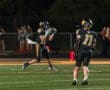 Quarterback #15 catches a football on a night field, with teammates and cheerleaders in the background.