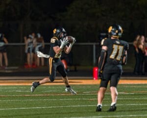 Quarterback #15 catches a football on a night field, with teammates and cheerleaders in the background.