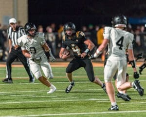 High school football player in black uniform evades a defender in white during a night game on a turf field.