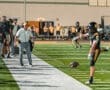 Football coach observing player during game practice, as teammates cheer from the sidelines under stadium lights.