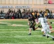 High school football player in black jersey evades defender while running with the ball on a field during a game.