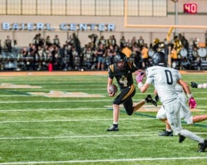 High school football player in black jersey evades defender while running with the ball on a field during a game.