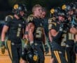 High school football players in black uniforms with paw print helmets, focused during a night game.