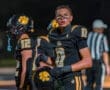 High school football player in black uniform, looking determined on the field during a night game with teammates nearby.