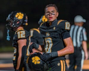 High school football player in black uniform, looking determined on the field during a night game with teammates nearby.
