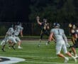 Quarterback in black jersey throwing a pass during a night football game against gray-clad defenders on the field.