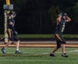 Two football players on the field, one celebrating and the other reacting, under bright stadium lights at night.