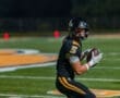 High school football player in black jersey prepares to throw a football on a well-lit field during a night game.