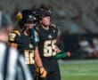 Two young football players in black jerseys on the sidelines, one holding a water bottle and focusing on the game.