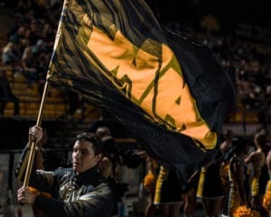 Cheerleader proudly waves a black and yellow school flag at a spirited event, surrounded by enthusiastic supporters.