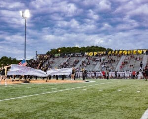 Cheerleaders run onto the field with an American flag at a North Allegheny football game under dramatic skies.