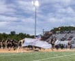 High school football team entering the field with cheerleaders, flags, and an American flag during a night game.