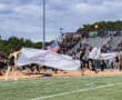Cheerleaders and football players burst onto the field, waving banners and a flag, during a spirited game kickoff.
