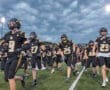High school football team running onto the field, wearing black and gold uniforms, under a dramatic sky.