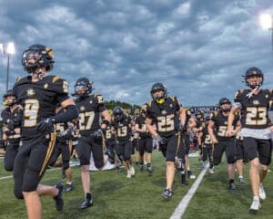 High school football team running onto the field, wearing black and gold uniforms, under a dramatic sky.