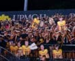 High school football fans in team colors cheer with large player cutouts at a night game event. Excitement fills the stands.