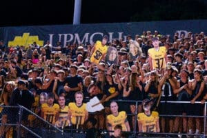 High school football fans in team colors cheer with large player cutouts at a night game event. Excitement fills the stands.