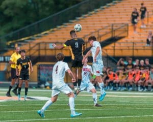 High school soccer match with players in action, one leaping to head the ball amid a vibrant crowd on the sidelines.