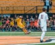 Youth soccer match, goalkeeper in yellow dives for the ball, while teammates and opponents watch from sidelines.
