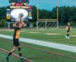 Young soccer player executing a throw-in on a field, with a scoreboard and teammate in the background.
