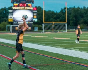 Young soccer player executing a throw-in on a field, with a scoreboard and teammate in the background.