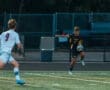 High school soccer player in a black jersey dribbles the ball past an opponent on the field during a competitive match.