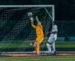 Goalkeeper in yellow leaping to block a soccer ball during a night match, with a player in white nearby.