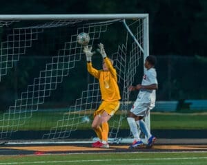 Goalkeeper in yellow leaping to block a soccer ball during a night match, with a player in white nearby.