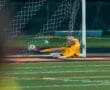 Goalkeeper in yellow gear dives for a soccer ball in the net, capturing an intense moment during the match.
