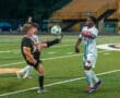 Two young soccer players in action, one juggling the ball while another defends during a night game on a green field.