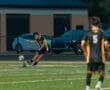 High school soccer player in black and yellow uniform kicking a ball during a match on a field at night.