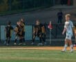 Soccer players celebrate a goal; teammates hug in joy while an opponent walks by near the corner flag.