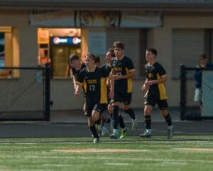 High school soccer players celebrating a goal under stadium lights, wearing black and yellow uniforms.