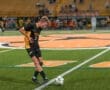High school soccer player in a black jersey dribbles the ball on a field with orange markings during a night game.