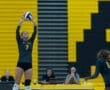 Volleyball player sets the ball in a gym, with teammates and commentators in the background. Bright yellow wall backdrop.