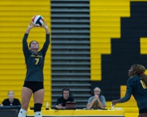 Volleyball player sets the ball in a gym, with teammates and commentators in the background. Bright yellow wall backdrop.