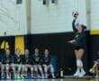 Volleyball player jumping to serve with teammates watching in the background during a game practice session.