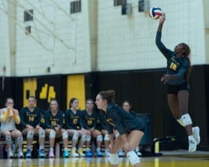 Volleyball player serves in a competitive match, teammates focused on gameplay in the background.