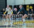 Two female volleyball players on the court; one prepares to receive a serve while the other watches closely.