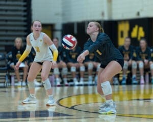 Two female volleyball players on the court; one prepares to receive a serve while the other watches closely.