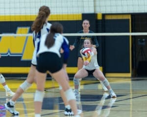 Volleyball players in a match, focusing on a player preparing to receive the ball at the net in a gym setting.