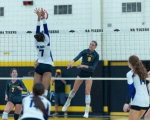 Two volleyball players leap at the net in an intense match, showcasing athleticism and teamwork in a gym setting.