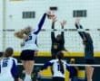 High school volleyball match in action, featuring players jumping to block a spike at the net.